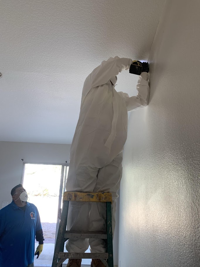 Technician in full protective suit drilling into ceiling during water damage inspection, supervised by another crew member in a mask